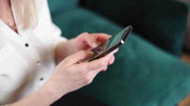 Evde akıllı telefon kullanarak kadın ellerini çıplak tırnak cilasıyla kapat. Yeşil kanepe arka planı. 