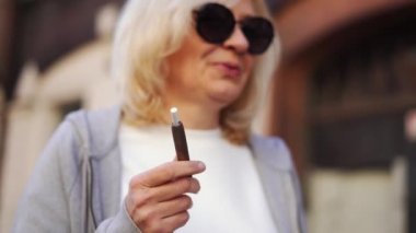 Orta yaşlı sarışın Avrupalı kadın sokakta sıcak tabacco e-sigara içiyor. Alternatif sigara içme yöntemi