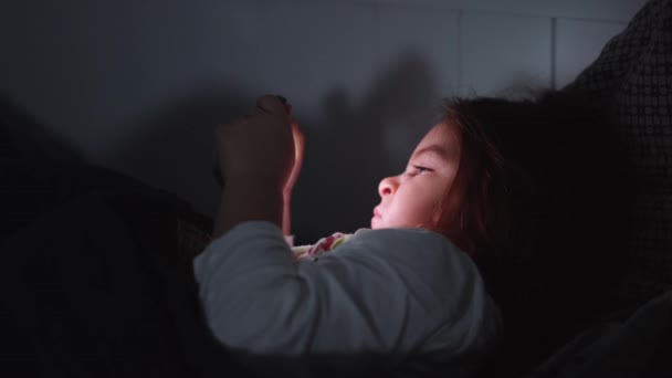 Dítě dívka se dívá na video na smartphone v posteli před spaním večer — Stock video