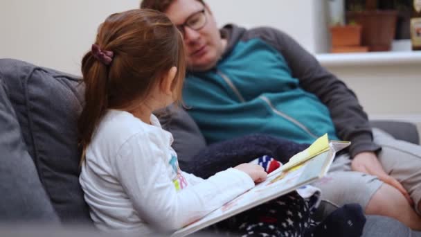 Ragazza caucasica prescolare con papà a casa a leggere libri sul divano prima di andare a letto. congedo di paternità, padre che si prende cura del bambino — Video Stock