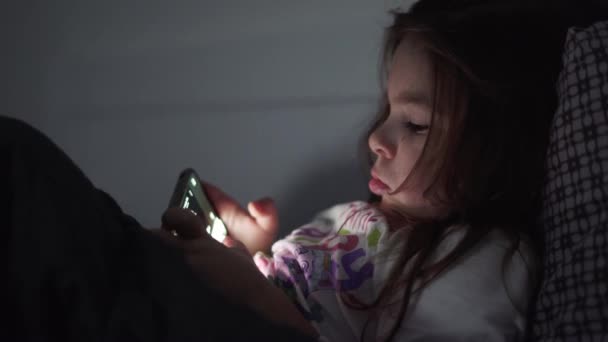 Dítě dívka se dívá na video na smartphone v posteli před spaním večer — Stock video