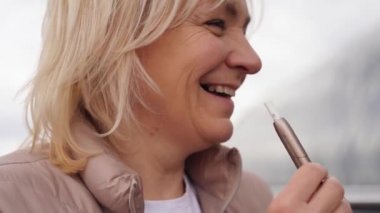 Orta yaşlı sarışın Avrupalı kadın sokakta sıcak tabacco e-sigara içiyor. Alternatif sigara içme yöntemi