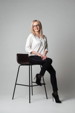 Gri arka planı olan bir stüdyoda, sandalyede oturan gözlüklü, gülümseyen genç bir kadın. Satış temsilcisi, yönetici, ajan, asistan, öğretmen, öğrenci