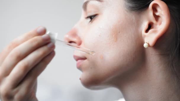 Close-up van vrouwelijke jonge blanke vrouw die serum of olie huidverzorgingsproduct op haar gezicht aanbrengt. Wellness en spa thuis — Stockvideo