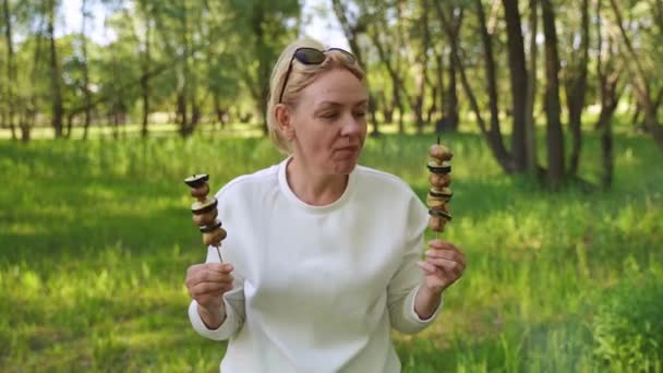 Mature woman 50 years old eating grilled vegetables from wooden skewers outdoors in forest — Stock Video