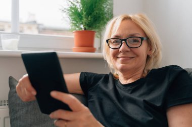 Olgun orta yaşlı bir kadın evinde, koltukta e-kitap satıyor. Yüksek kalite fotoğraf