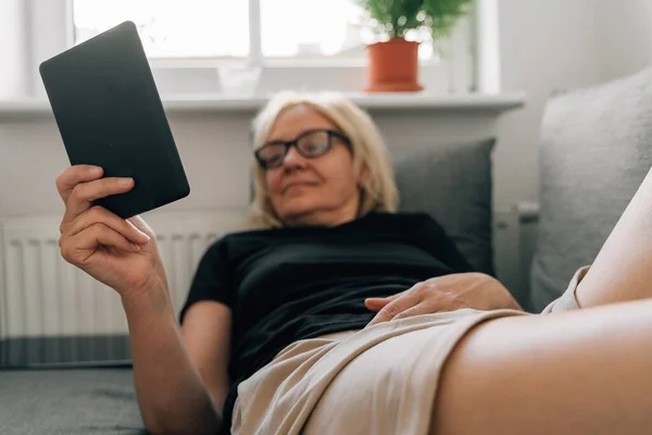 一个成熟的中年女人在家里沙发上卖电子书 高质量的照片 — 图库照片