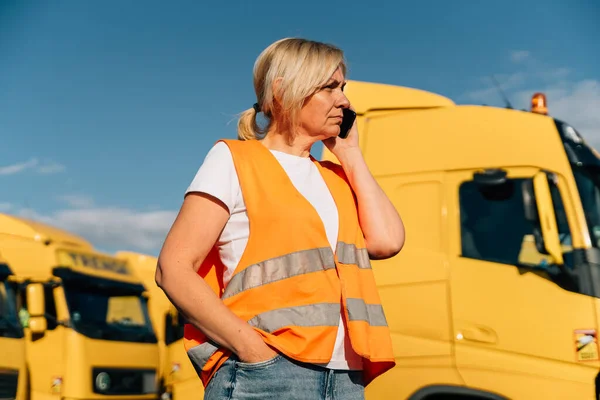 Happy middle-aged woman having a phone call in front of yellow semi-truck vehicle . High quality photo