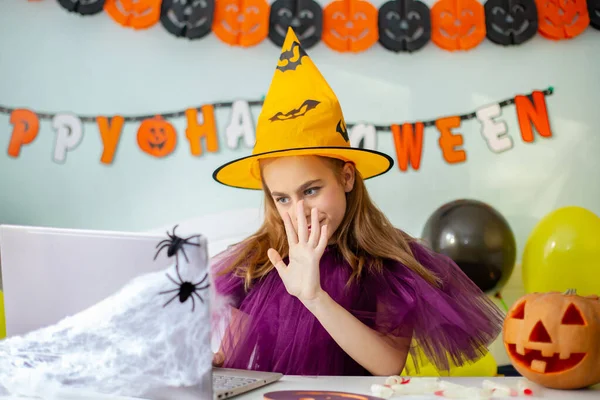 Cute little girl wearing witch hat sitting behind a table in Halloween theme decorated room. Halloween party concept. Preparation for the celebration, internet communication.