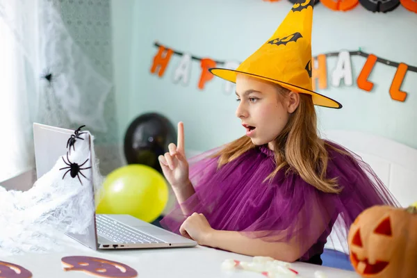Cute little girl wearing witch hat sitting behind a table in Halloween theme decorated room. Halloween party concept. Preparation for the celebration, internet communication.