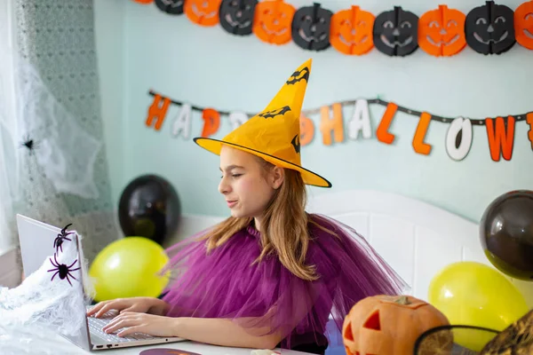 Cute little girl wearing witch hat sitting behind a table in Halloween theme decorated room. Halloween party concept. Preparation for the celebration, internet communication.