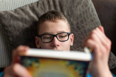 Kanepede kitap okuyan gözlüklü çocuk.