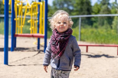 Güneşli bir günde, bir çocuk parkında dikilen atkı ve bulanık arkaplanı olan kazak giyen sevimli çocuğun ön portresini kapat ve boşluğu kopyala..