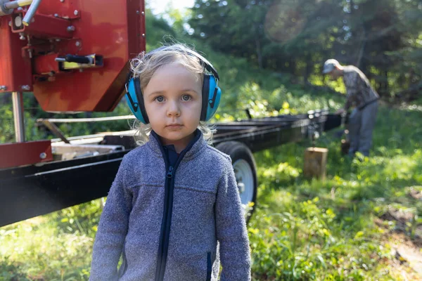 Close up front portrait of a cute three year old boy wearing ear defenders, as a blurry lumberjack is seen in background setting up industrial saw.