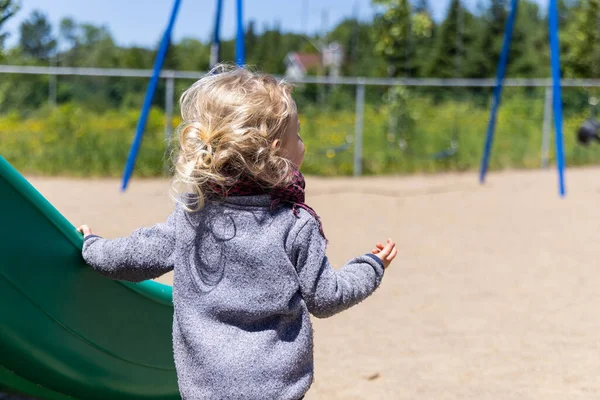 A two year old boy with wavy blonde hair is seen wearing grey jumper with back to camera, standing by a slide on a play park with copy space to right.