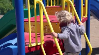 Camera panning around the rear of a two year old boy with blonde wavy hair playing at a recreational park, climbing the steps to a slippery slide.