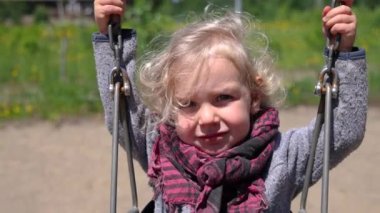Closeup slow motion clip of an adorable three year old caucasian boy, sitting in swing and holding on to chains at a playground with blurry background.