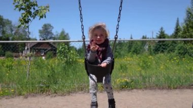 Slow motion clip of a cute two year old boy seated in a toddler swing at a playground, facing the camera and waving hand hello. With copy space.