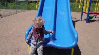 Slow mo clip of a happy 2 year old caucasian boy wearing red checkered scarf, sliding down a slippery slope then runs to steps for another go at park.
