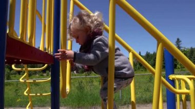 Closeup side view in slow motion as two year old boy wearing red scarf, cautiously steps on bars of a climbing frame and play set at local playground.