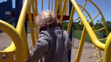 Slow motion video from behind a daring 3 year old toddler with blonde wavy hair, climbing up the yellow bars of a playground slide set at local park.