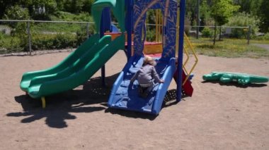Slow motion camera panning on a carefree young toddler, with wavy blonde hair kneeling down to climb up a playground climbing frame and slide set.