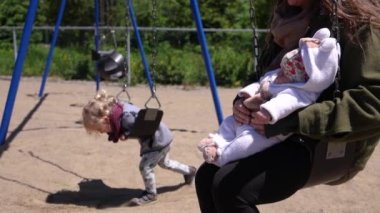 Closeup selective focus video of a mother sitting on a swing cradling newborn baby girl, blurry toddler is seen swaying back and forth in background.