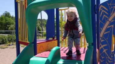 An adventurous two year old boy is seen with enthusiastic facial expression at the top of a playground slide before sliding down towards camera.