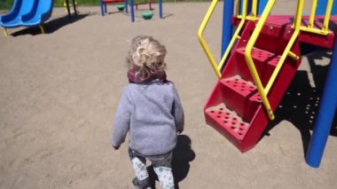 Arkasından kumlu zeminde zıplayan ve çocuk parkındaki kaydırağına doğru koşan heyecanlı iki yaşındaki bir çocuğun yavaş çekim videosu..