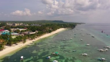 Drone footage along a stretch of coastline in the Philippines with beachfront hotels sticking above palm trees and traditional boats in shallow sea.