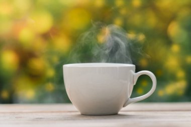 Ahşap zeminde beyaz seramik kahve kupası, yeşil ağaç bokeh arka plan. Yumuşak odaklanma. Sığ odak etkisi.