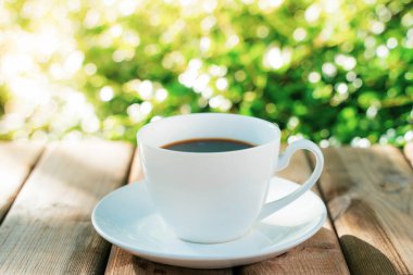 Ahşap zeminde beyaz seramik kahve kupası, yeşil ağaç bokeh arka plan. Yumuşak odaklanma. Sığ odak etkisi.