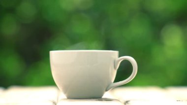 Ahşap zeminde beyaz seramik kahve kupası, yeşil ağaç bokeh arka plan. Yumuşak odaklanma. Sığ odak etkisi.
