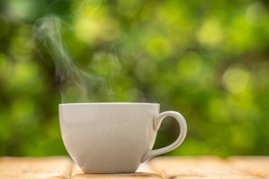 Ahşap zeminde beyaz seramik kahve kupası, yeşil ağaç bokeh arka plan. Yumuşak odaklanma. Sığ odak etkisi.