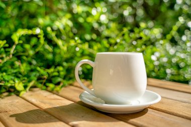 Ahşap zeminde beyaz seramik kahve kupası, yeşil ağaç bokeh arka plan. Yumuşak odaklanma. Sığ odak etkisi.