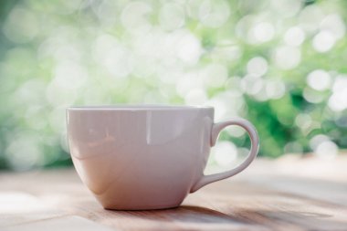 Ahşap zeminde beyaz seramik kahve kupası, yeşil ağaç bokeh arka plan. Yumuşak odaklanma. Sığ odak etkisi.