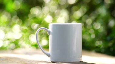 Ahşap zeminde beyaz seramik kahve kupası, yeşil ağaç bokeh arka plan. Yumuşak odaklanma. Sığ odak etkisi.