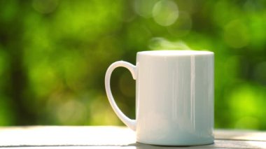 Ahşap zeminde beyaz seramik kahve kupası, yeşil ağaç bokeh arka plan. Yumuşak odaklanma. Sığ odak etkisi.