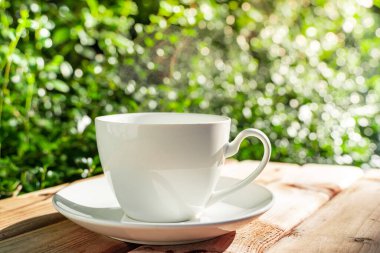 Ahşap zeminde beyaz seramik kahve fincanı, bahçede sabah güneşinde yeşil ağaç bokeh arka plan. Yumuşak odaklanma. Sığ odak etkisi.
