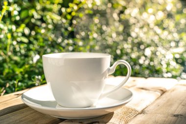 Ahşap zeminde beyaz seramik kahve fincanı, bahçede sabah güneşinde yeşil ağaç bokeh arka plan. Yumuşak odaklanma. Sığ odak etkisi.