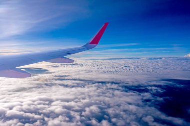 Uçuş sırasında uçağın kanatları yolcu penceresinden bakar. Mavi gökyüzünde beyaz bulutlar var.