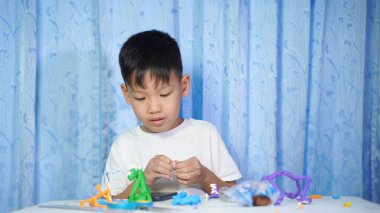 Konsantre olmuş Asyalı çocuk evdeki beyaz bir masada oturup plastik bebeklerden yapıyor.