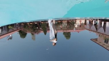 Siyah mayo giymiş genç bir kadın. Beyaz, uzun pelerinli. Açık havuza dalıyor ve mavi fayanslarla süsleniyor..