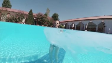 Siyah mayo giymiş genç bir kadın ve açık havuzun dibinde mavi fayanslarla süslenmiş beyaz bir pelerin duruyor..