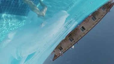 Siyah mayo giymiş genç bir kadın ve açık havuzun dibinde mavi fayanslarla süslenmiş beyaz bir pelerin duruyor..