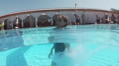 Siyah mayo giymiş genç bir kadın ve açık havuzun dibinde mavi fayanslarla süslenmiş beyaz bir pelerin duruyor..