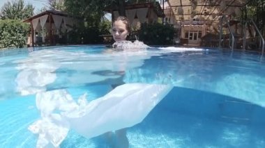 Siyah mayo giymiş genç bir kadın ve açık havuzun dibinde mavi fayanslarla süslenmiş beyaz bir pelerin duruyor..