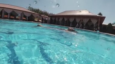 Siyah mayo giymiş genç bir kadın ve açık havada yüzme havuzunda mavi fayanslarla süslenmiş beyaz bir pelerin yüzüyor..