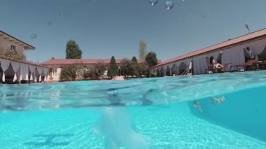 Siyah mayo giymiş genç bir kadın ve açık havada yüzme havuzunda mavi fayanslarla süslenmiş beyaz bir pelerin yüzüyor..