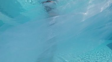 Siyah mayo giymiş genç bir kadın ve açık havada yüzme havuzunda mavi fayanslarla süslenmiş beyaz bir pelerin yüzüyor..